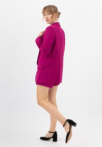 Blazer fucsia con rever e due tasche laterali, abbinato a pantaloni shorts coordinati. Scarpe con tacco e cinturino alla caviglia nere completano l'outfit.