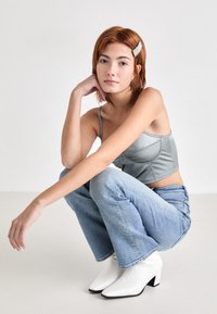 Top corto grigio con spalline sottili, jeans flared azzurri e stivaletti bianchi con tacco. Modella in posa accovacciata con una clip per capelli.
