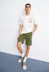 White t-shirt with a colorful print and "ELLESSE" logo, paired with olive green shorts featuring zip pockets. Beige sneakers with green accents.