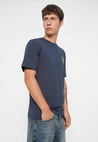 Marineblå bomulds t-shirt med rund hals og korte ærmer; har et lille gult grafisk motiv på brystet. Båret med denimjeans.