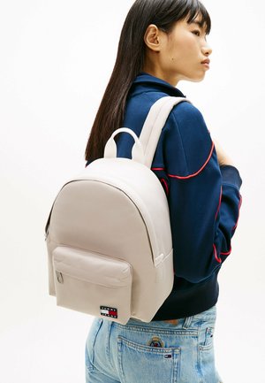 Vrouw met een marineblauw jack en lichtblauwe spijkerbroek, die een beige Tommy Jeans-rugzak draagt, kijkend naar rechts tegen een effen witte achtergrond.