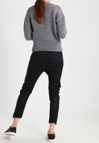 Šedý pletený svetr s ribovaným lemem, v kombinaci s černými úzkými džíny. Oblečení doplňují černé nazouvací boty, pohled zezadu.