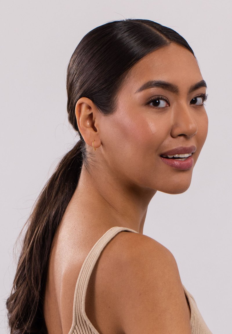 Goldene Creolen an einem Model mit schulterlangem, mittelbraunem Haar, das ein hellbeiges, geripptes Oberteil trägt. Glatte Haut mit natürlichem Makeup, leicht nach links gewandt.