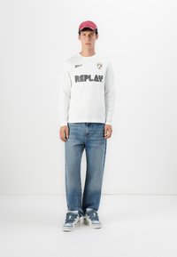 Ung mand står op, iført hvid langærmet skjorte med "REPLAY" logo, blå jeans, blå og hvide sneakers og rød kasket mod en hvid baggrund.