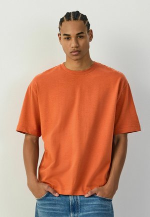 Young man with braided hair wearing an oversized orange T-shirt and blue jeans, standing with hands in pockets against a plain background.