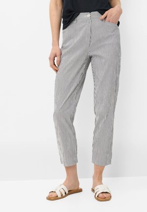 Persoon die cropped, slim-fit broek met verticale zwart-witte strepen draagt, een zwarte top en witte slippers, staand tegen een witte achtergrond.