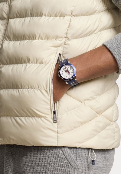 Gilet matelassé beige avec une fermeture éclair argentée, texture de pull gris, et une montre bleu marine avec un cadran blanc et des détails bleus sur un bracelet sombre.