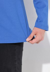 Blaues Langarmhemd aus Baumwolle mit geradem Saum. Eine Hand zieht am unteren Rand des Hemdes. Dunkler Stoff ist darunter sichtbar.