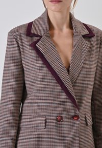 Bruin en beige geruit blazer met een diep paarse fluwelen reven, dubbele knopen en zijzakken, met gestructureerde schouders.