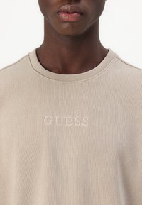 Béžová žíhaná mikina s kulatým výstřihem, která má vyšívané logo "GUESS" v světlé nitě. Tkanina vypadá měkce a má texturu.
