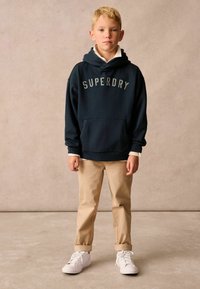 Sudadera azul marino con el logo "SUPERDRY", bolsillo canguro; combinada con chinos beige y zapatillas blancas. El modelo está de pie frente a un fondo neutro.