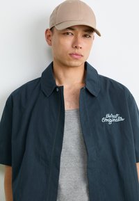 Mann som har på seg en beige caps, mørk marineblå jakke med teksten "the New Originals", og en grå tank top, står mot en enkel hvit bakgrunn.