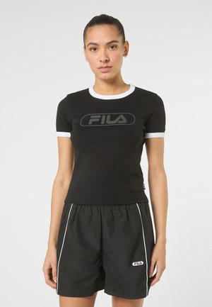 Młoda kobieta związała włosy do tyłu, ma na sobie czarną koszulkę FILA z białą lamówką i czarne spodenki FILA z białym paskiem, stoi na tle jednolitego tła.