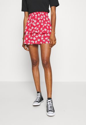 Vrouw die een rode mini-rok met tiers en madeliefjesprint, een zwart T-shirt en zwarte hoge sneakers met witte veters draagt, staand op een grijze vloer.