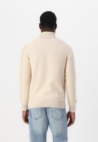 Maglione beige lavorato a maglia con collo alto a coste e polsi a coste, abbinato a jeans in denim azzurro chiaro. La texture è morbida e liscia.