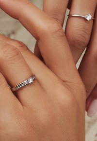 Deux anneaux en argent sur des mains ; l'un présente une pierre centrale claire avec de petites pierres d'accent, l'autre a une seule pierre claire. Texture lisse.