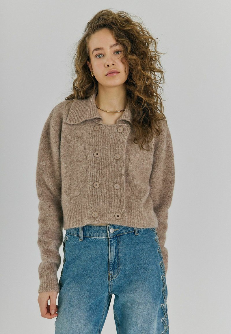 Brauner, wolliger, kurzer Pullover mit einem gerippten Kragen, doppelt geknöpften Knöpfen und langen Ärmeln, kombiniert mit hoch taillierten blauen Jeans.