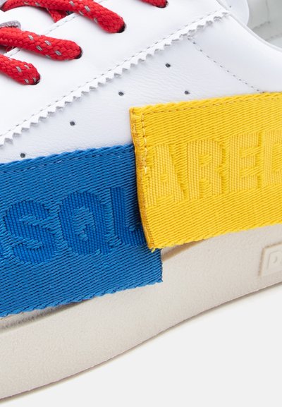 Λευκό αθλητικό παπούτσι με κόκκινα κορδόνια, μπλε και κίτρινες υφασμάτινες λωρίδες με ανάγλυφ η γραφή. Στρογγυλεμένη μύτη και υφή στη σόλα.