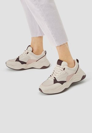 Des baskets présentant un mélange de matériaux beige, rose clair et marron foncé. Semelle épaisse, panneaux texturés et accents contrastants sont bien visibles.