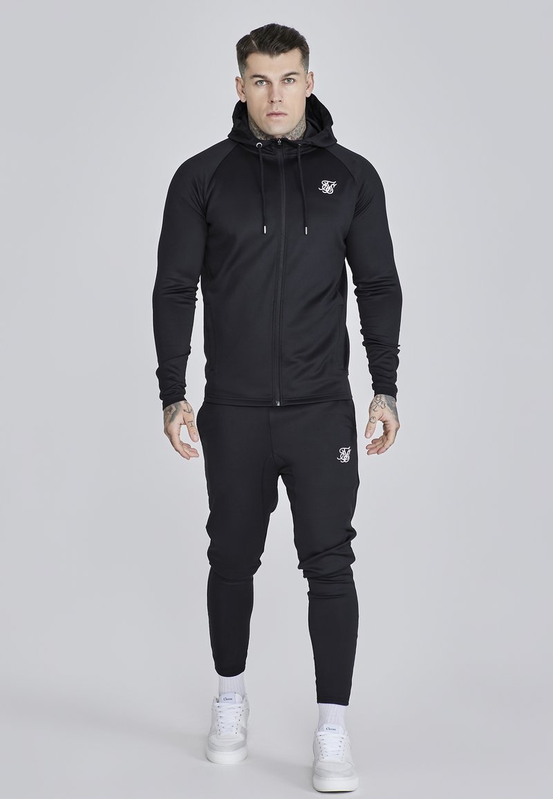 Man met tatoeages die een zwarte zip-up hoodie en bijpassende joggingbroek met witte sneakers draagt, staand tegen een grijze achtergrond.