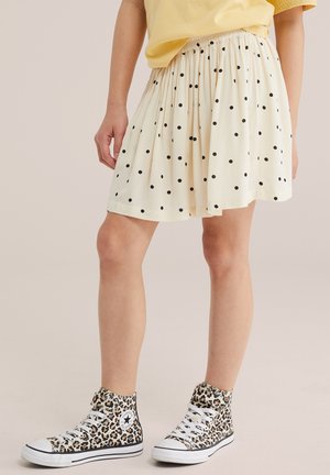 Osoba nosiaca krémovú sukňu s čiernymi bodkami, žltý top a vysoké tenisky so vzorom leopard s bielymi šnúrkami.
