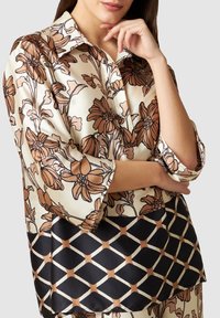 Blusa con motivo floreale nei colori marrone e crema, finitura satinata, maniche a tre quarti e un motivo geometrico nero lungo l'orlo.
