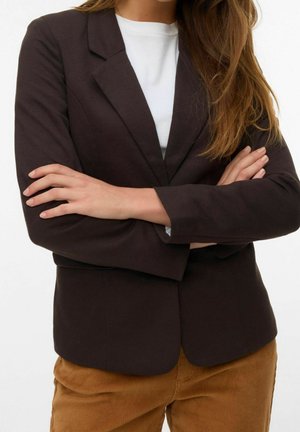 Frau mit langen Haaren, die einen dunkelbraunen Blazer, ein weißes Top und hellbraune Hosen trägt, mit verschränkten Armen.