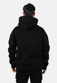 Sudadera con capucha negra que presenta una capucha con cordón, un ajuste holgado y puños acanalados, mostrada desde atrás. El material parece suave y resistente.