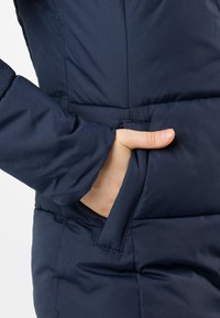 Navy puffer jacket detail showing a hand in a side pocket with a fitted sleeve, featuring quilted texture and smooth fabric.