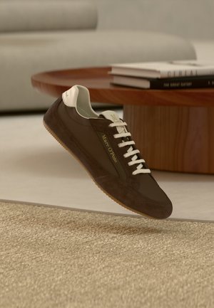 Brauner und beiger Marc O'Polo-Sneaker mit weißen Schnürsenkeln, der über einem strukturierten beigen Teppich in einem Wohnzimmer schwebt.