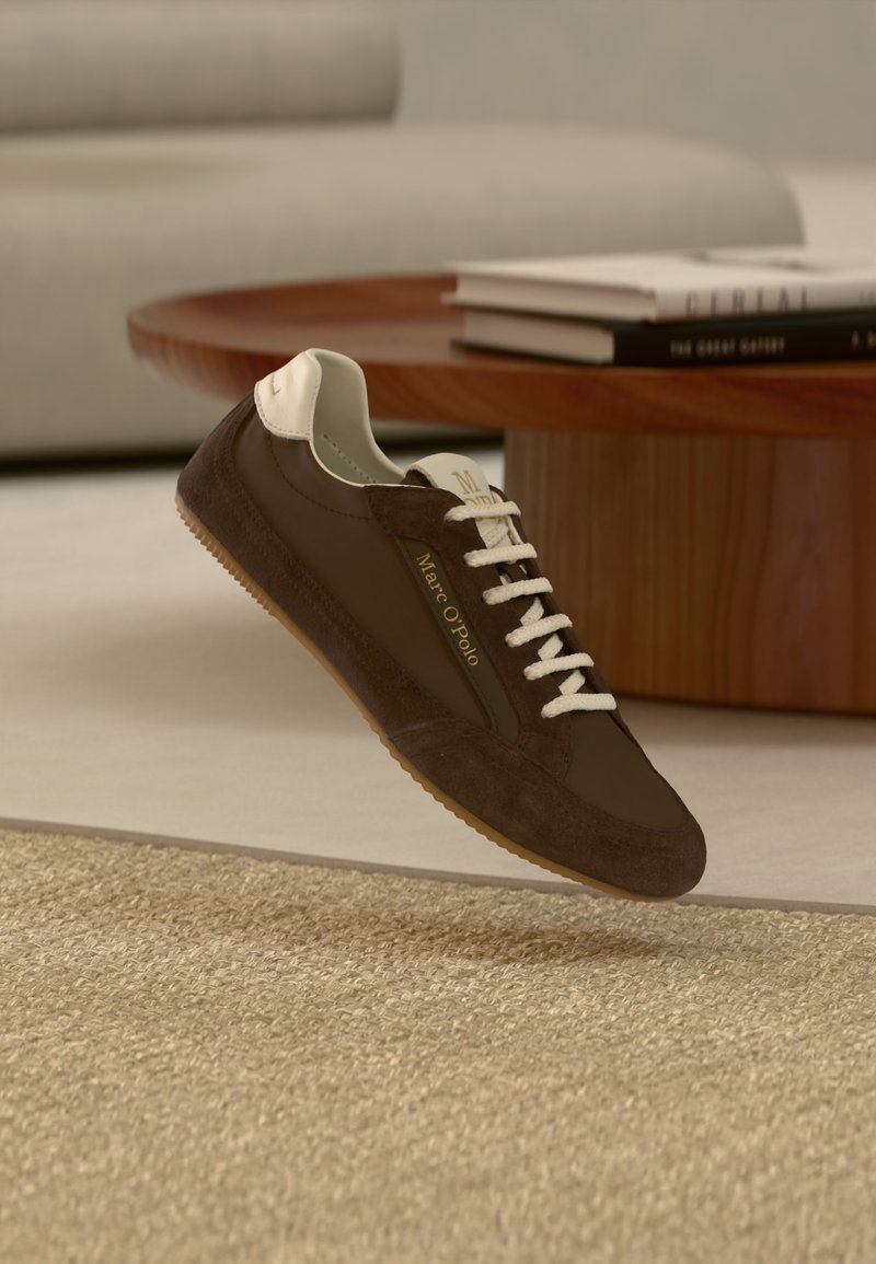 Καφέ και μπεζ αθλητικό παπούτσι Marc O'Polo με λευκά κορδόνια που αιωρείται πάνω από μια υφασμάτινη μπεζ μοκέτα σε ένα καθιστικό.
