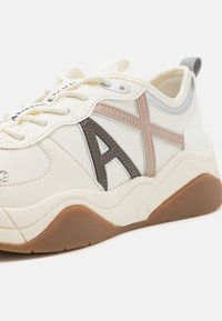 Sneaker color crema caratterizzata da un mix di materiali sintetici, logo prominente e accenti in marrone e beige contrastanti su una suola testurizzata.