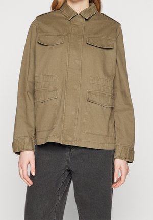 Femme portant une veste de style militaire kaki avec des poches à rabat et un jean noir, debout devant un fond uni.