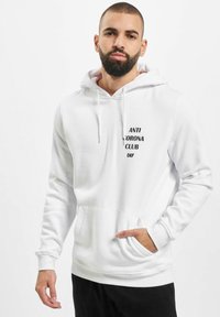 Weißer Kapuzenpullover aus weichem Material mit einer vorderen Kängurutasche. Schwarzer Drucktext "ANTI CORONA CLUB DEF" auf der Brust.