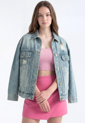 Ung kvinde iført en lyseblå slidt denimjakke over en pink kort top og en pink nederdel, står med foldede hænder.