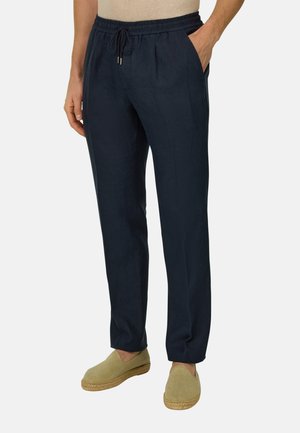 Homme portant un pantalon bleu marine à taille élastique avec cordon de serrage et des chaussures beige à enfiler, debout avec une main dans la poche.