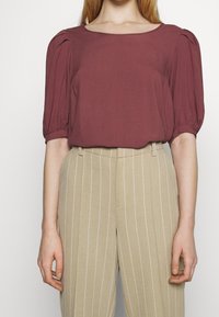 Bordowa bluzka z krótkim rękawem i kwadratowym dekoltem, z marszczonymi bufkami na rękawach. Połączona z beżowymi, wąsko prążkowanymi spodnie z wysokim stanem.