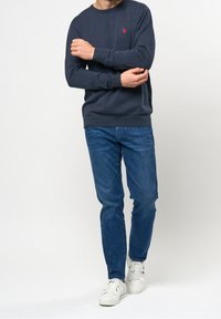 Man som bär en marinblå sweatshirt, blå jeans och vita sneakers, står mot en vit bakgrund och justerar ärmen med korsade armar.