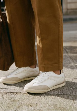 Henkilö, jolla on valkoiset ja beige tennarit ja ruskeat housut, seisoo auringonpaisteessa olevalla kivipäällysteellä ja pitää ruskeaa laukkua.