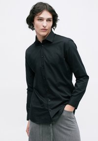 Cămașă neagră cu mâneci lungi, cu design ajustat, din țesătură de bumbac, textură fină, cu guler ascuțit și un discret rând de nasturi pe față.