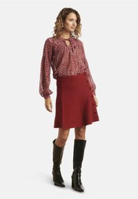 Red wool skirt with side buttons paired with a patterned burgundy and white blouse. Model wears tall black leather heeled boots.