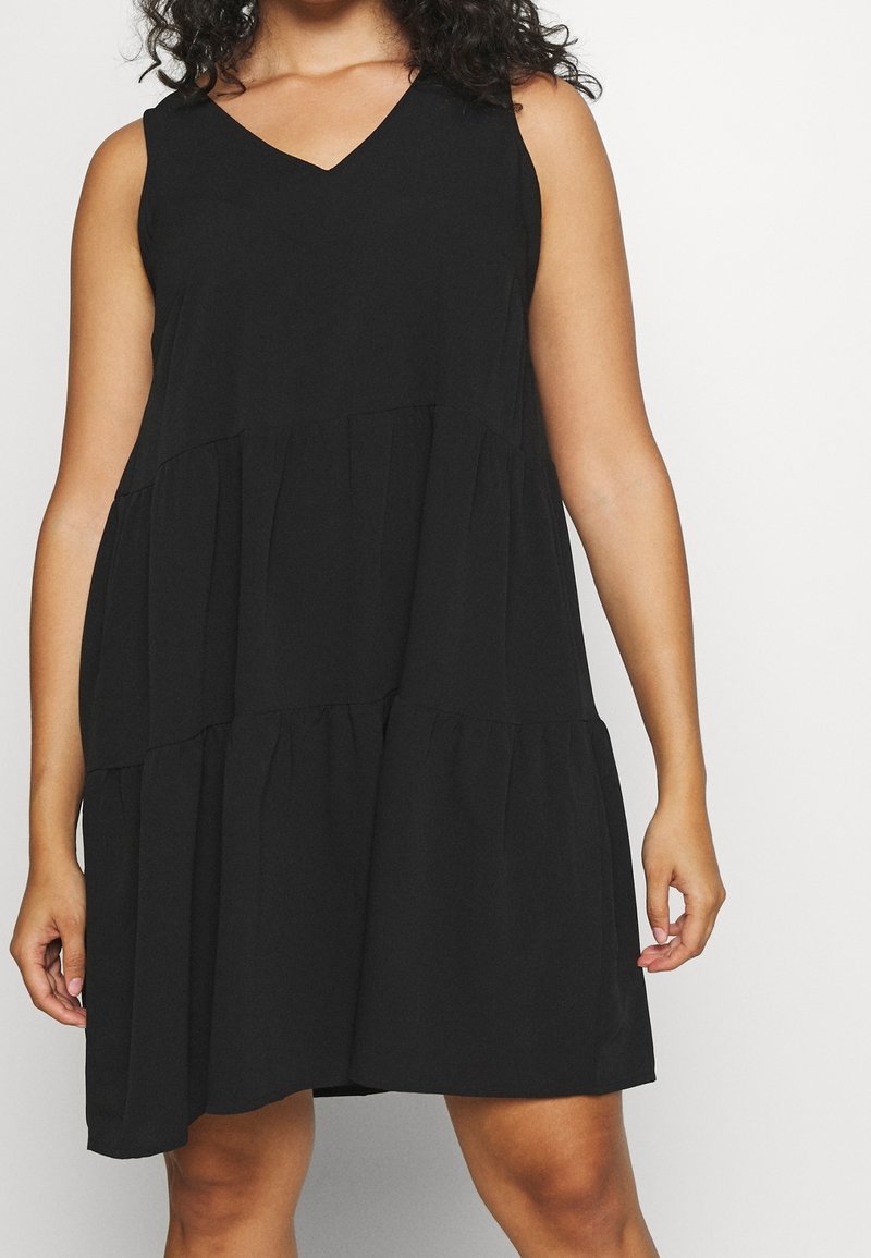 Femme portant une robe noire sans manches à volants avec un décolleté en V, debout devant un fond clair uni.