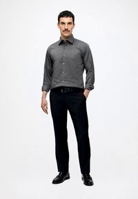 Bărbat cu păr închis la culoare și mustață, purtând o cămașă gri cu nasturi, pantaloni negri, curea neagră și pantofi negri, stând cu o mână în buzunar.