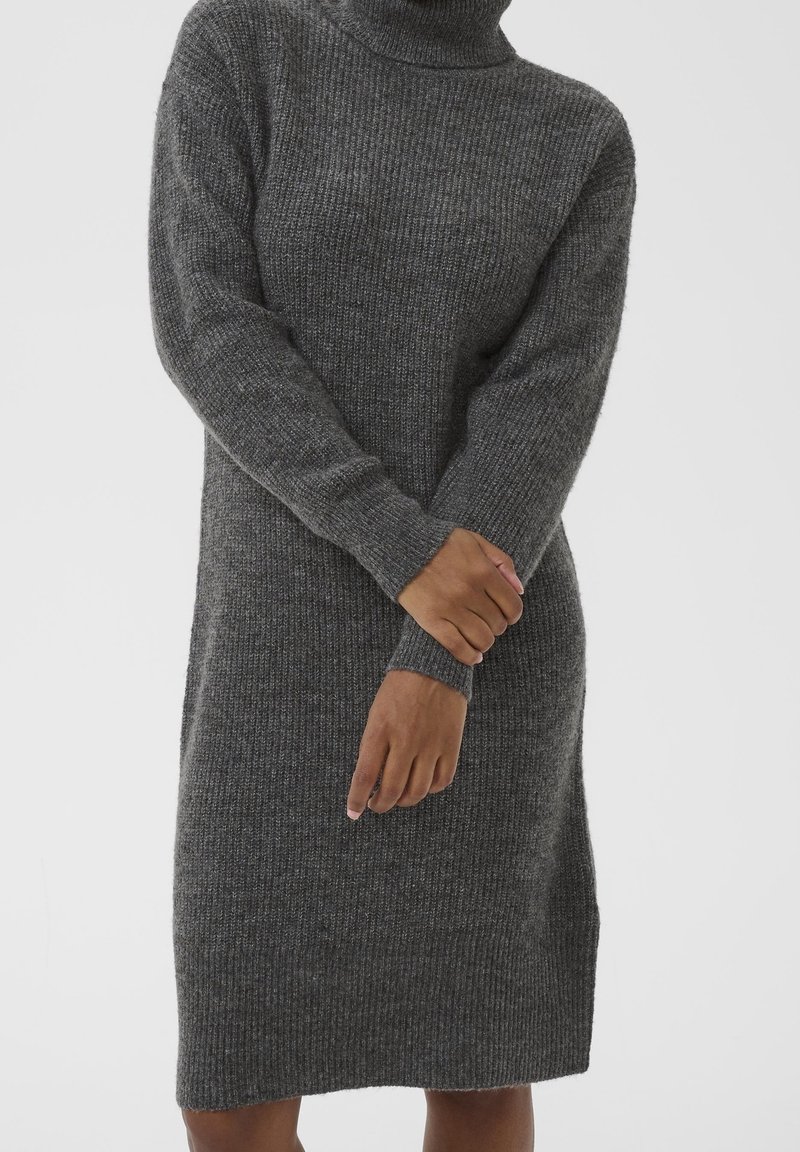 Personne portant une robe en tricot côtelé gris foncé à col roulé avec des manches longues, les mains croisées aux poignets devant.