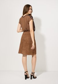 Braunes Kurzarmoberteil mit Tiermuster-Akzenten, kombiniert mit einem knie-langen Rock aus braunem Wildleder. Schwarze High-Heels vervollständigen das Outfit.