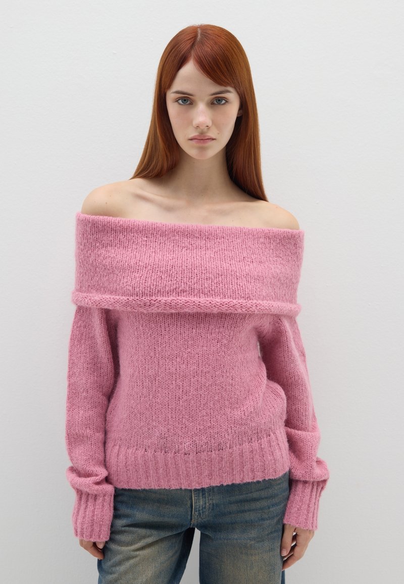 Rosa Off-Shoulder-Pullover aus weichem, strukturiertem Strickstoff. Gerippter Saum und Ärmelabschlüsse. Kombiniert mit hellblauen Jeans.