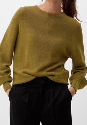 Mujer que lleva un suéter de punto verde oliva con mangas largas metido en unos pantalones negros de talle alto, con las manos en los bolsillos, sobre un fondo liso.