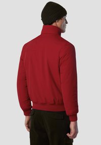 Giacca rossa imbottita con un alto colletto e polsini a coste. Texture liscia e design aderente con un orlo che si ferma in vita, indossata con pantaloni cargo color oliva.