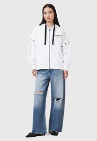 Sudadera blanca con cremallera y cordones negros, con un logo y detalles de broche. Combinada con unos vaqueros anchos desgastados de color azul claro.
