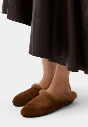 Pantofole in suede marrone con bordo in pelliccia beige morbida, caratterizzate da una punta rotonda e un design aperto sul retro, posizionate sotto una gonna in pelle marrone.